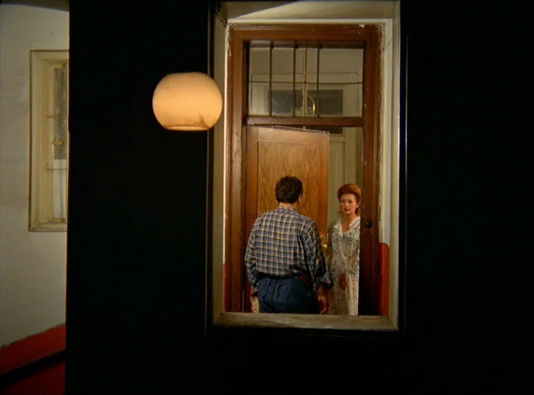 The Merchant Of The Four Seasons (1971) movie still: wide — A dark hallway with a white wall and red trim leads to an open wooden door. A man stands…