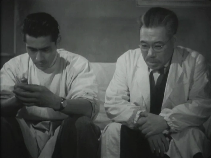 The Quiet Duel (1949) movie still: medium — A spare room features blank walls behind two men dressed in white medical coats. They sit close together…