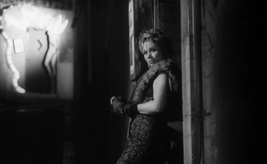 Vivement Dimanche (1983) movie still: medium — A dark city street at night with stone textures and a glowing neon sign. A woman in a fur wrap and…