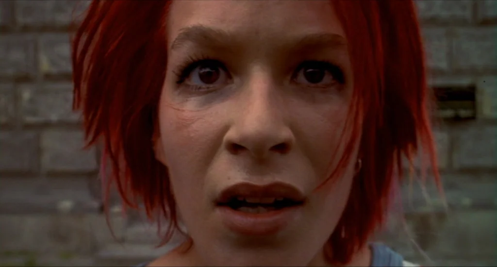Run Lola Run (1998) movie still: closeup — A close-up of a young woman with bright red hair against a weathered brick background. The subject gazes…