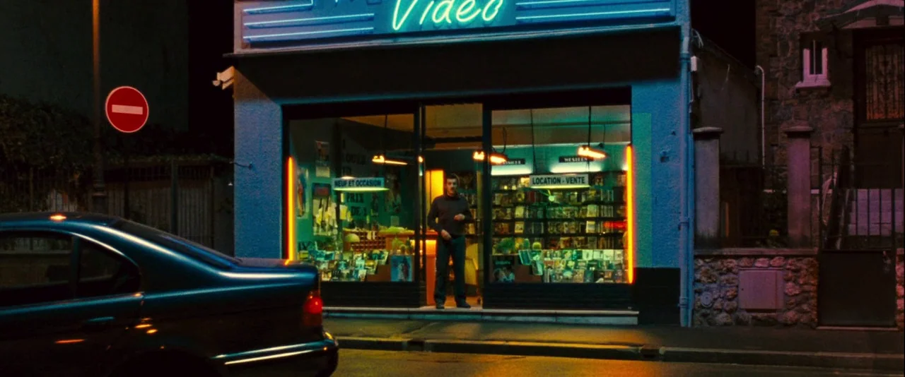 Micmacs (2009) movie still: wide — A nighttime storefront displays neon blue signage above a glass facade filled with media. A man stands motionless…