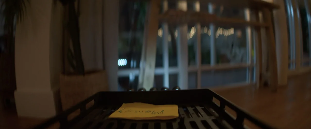 Drop (2025) movie still: closeup — A close-up view of a yellow note sitting on a dark metal frame. The blurred background shows a window with dark…
