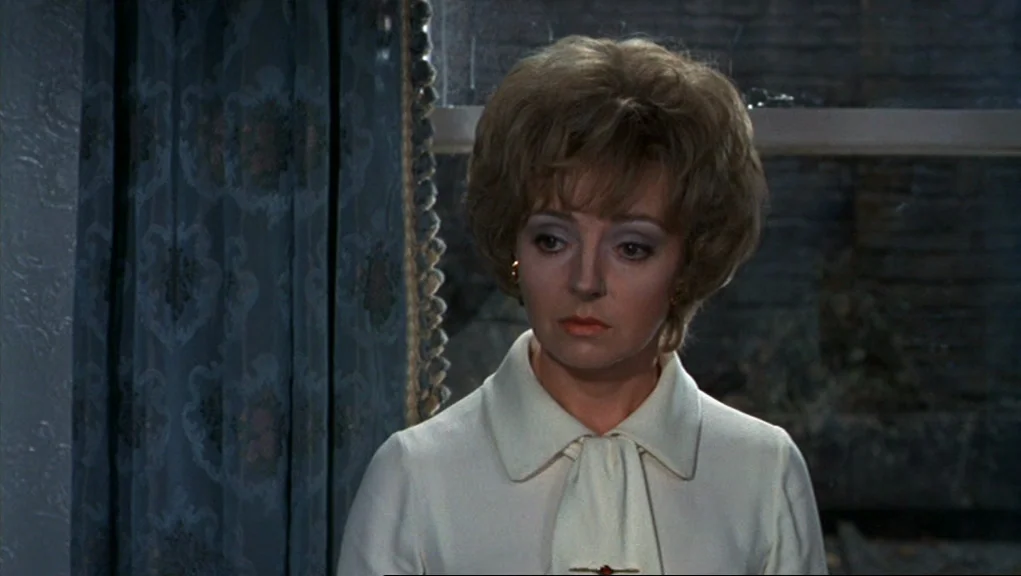 Frenzy (1972) movie still: medium — A woman stands indoors in front of a window with heavy blue patterned curtains. She looks downward while…