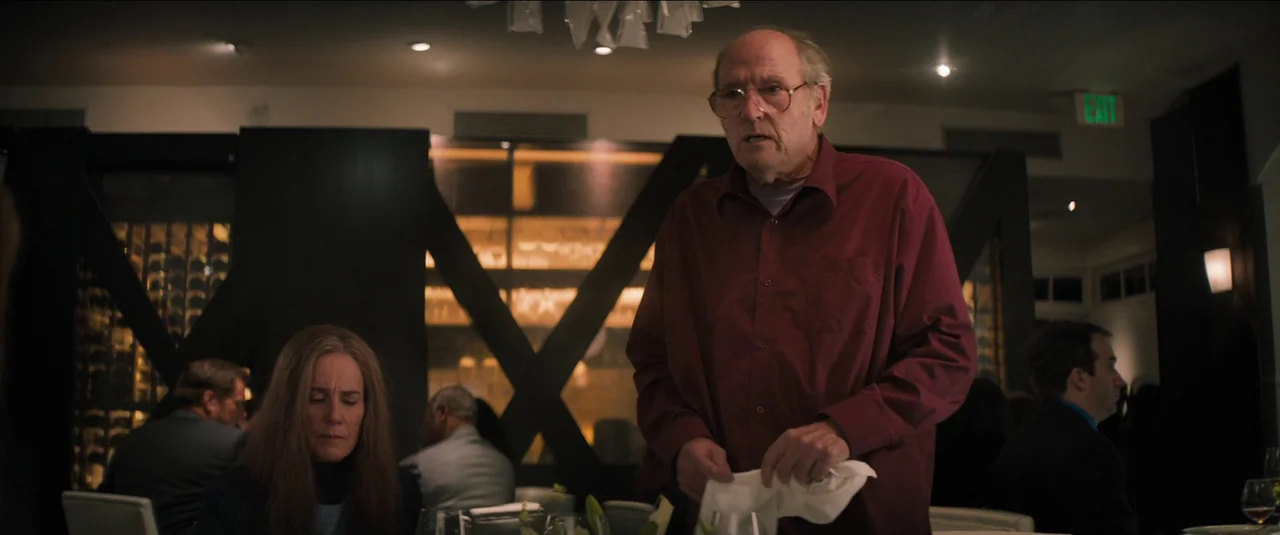 Kajillionaire (2020) movie still: medium — An upscale dining space features dark wood accents and a lit glass wine display. A man stands at a table…