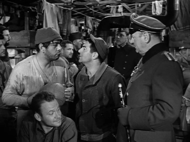Stalag 17 (1953) movie still: medium — A crowded wooden barracks features exposed beams and clothing hanging from rafters. Several men in varied…