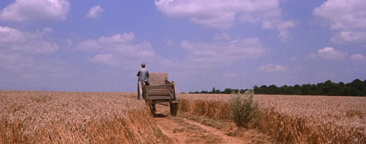 Lust For Life (1956) movie still: wide — A wide wheat field stretches under a expansive blue sky with scattered clouds. A man sits on a wooden cart…