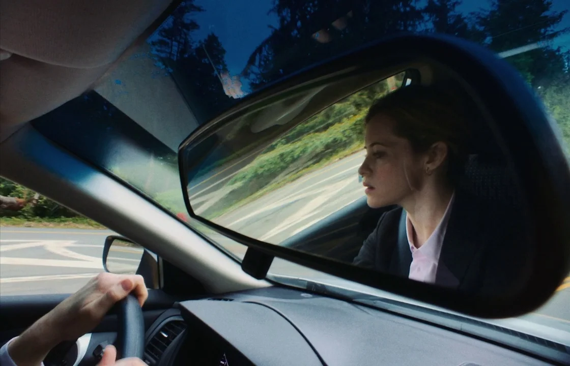 Unsane (2018) movie still: closeup — A woman's face is reflected in the rearview mirror of a car moving along. The mirror shows the profile of a…