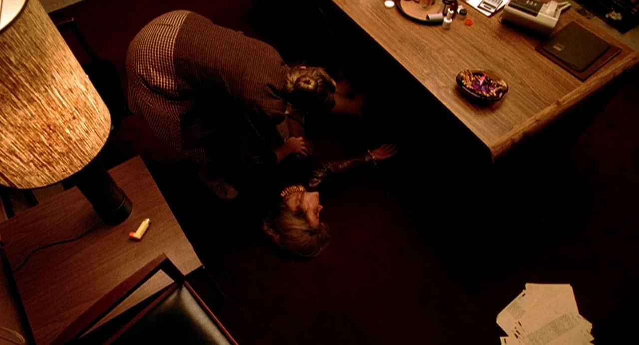 Office Killer (1997) movie still: wide — A dark, wood-paneled office contains a heavy desk and a floor lamp. One woman kneels over another woman who…