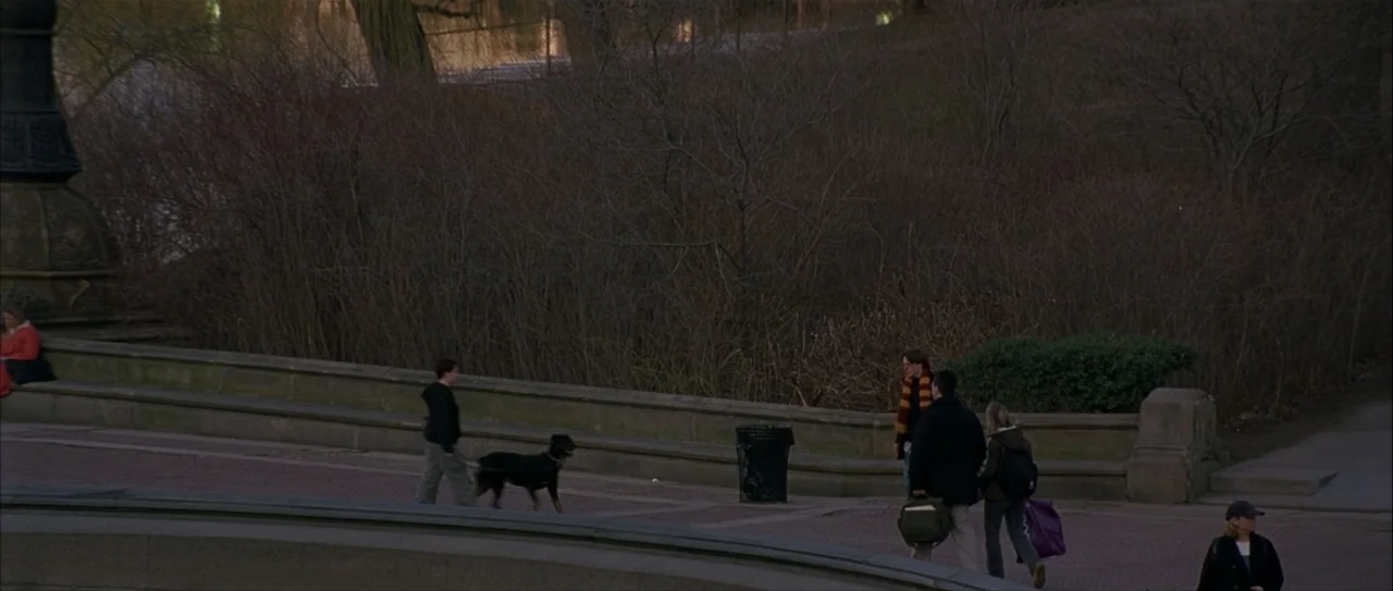 Igby Goes Down (2002) movie still: wide — A public walkway in a park features a low stone retaining wall and bare trees. Several pedestrians walk…