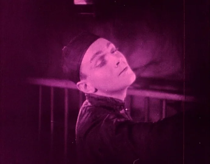 Broken Blossoms (1919) movie still: closeup — A person stands in a dim room with a visible railing behind them. The individual wears a dark cap and…