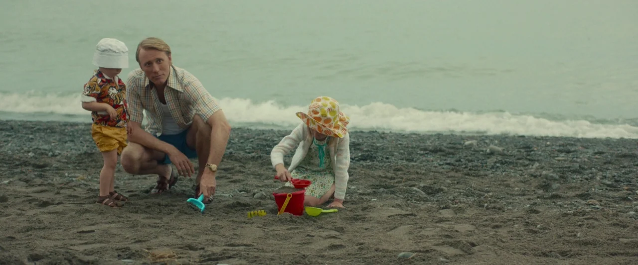 Stockholm (2019) movie still: wide — A grey pebble beach with gentle waves washing against the shore under overcast skies. A father crouches in the…