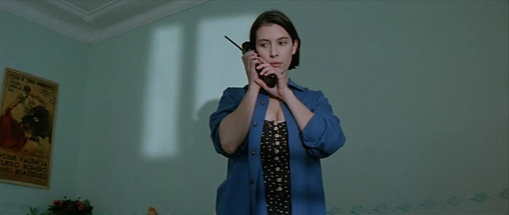 IP5 (1992) movie still: medium — A room with pale green walls and a bullfighting poster hung on the left. A young woman stands while holding a…