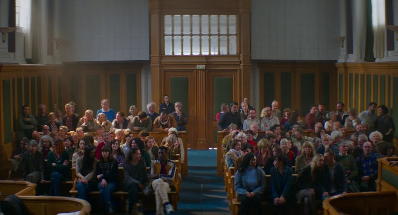 Anatomy of a Fall (2023) movie still: wide — A large courtroom filled with a crowd of people seated on wooden benches. The room features high…