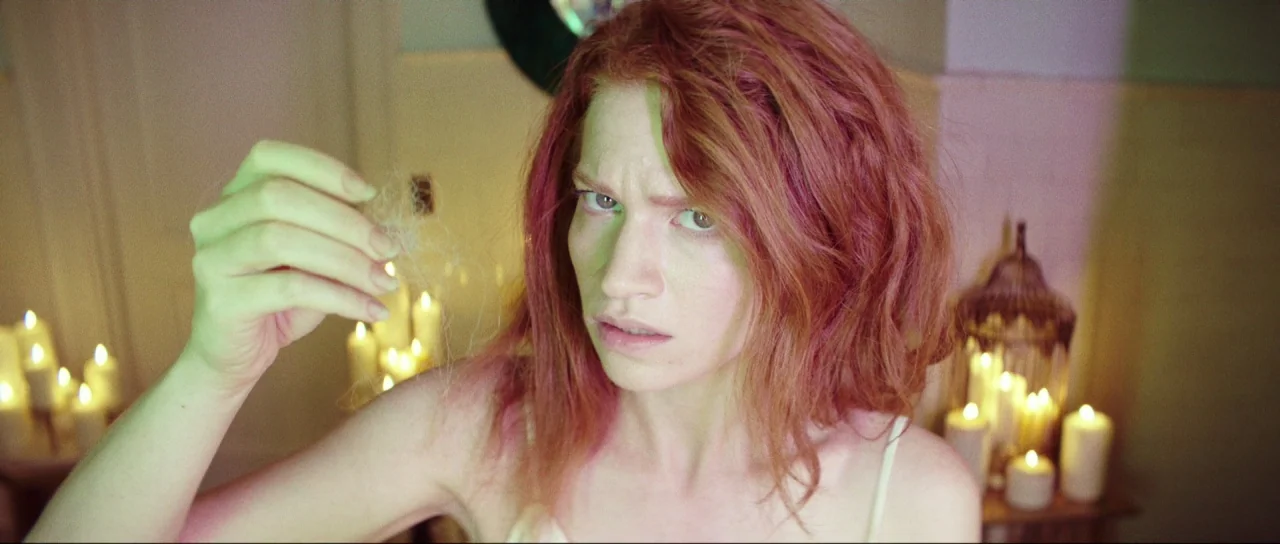 Braid (2018) movie still: closeup — A dimly lit interior space is crowded with multiple lit white candles. A young woman with pink-dyed hair looks…
