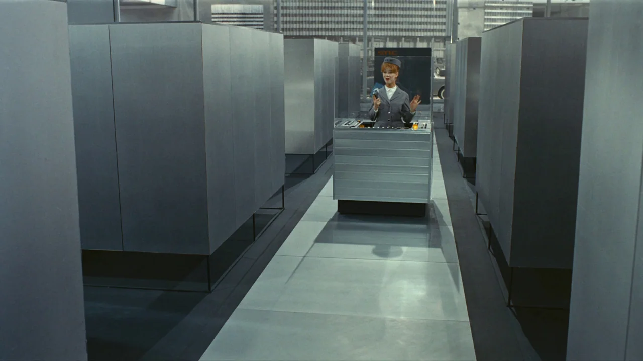Playtime (1967) movie still: wide — A spacious office interior features rows of repetitive metallic grey modular workstations. A young woman in a…