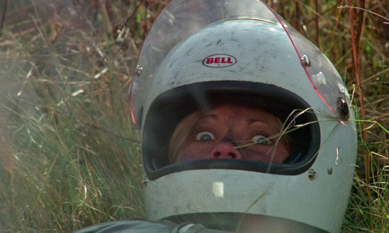 Rabid (1977) movie still: closeup — A person lies amidst dry, unkempt grass in an outdoor field. The individual wears a white helmet while staring…
