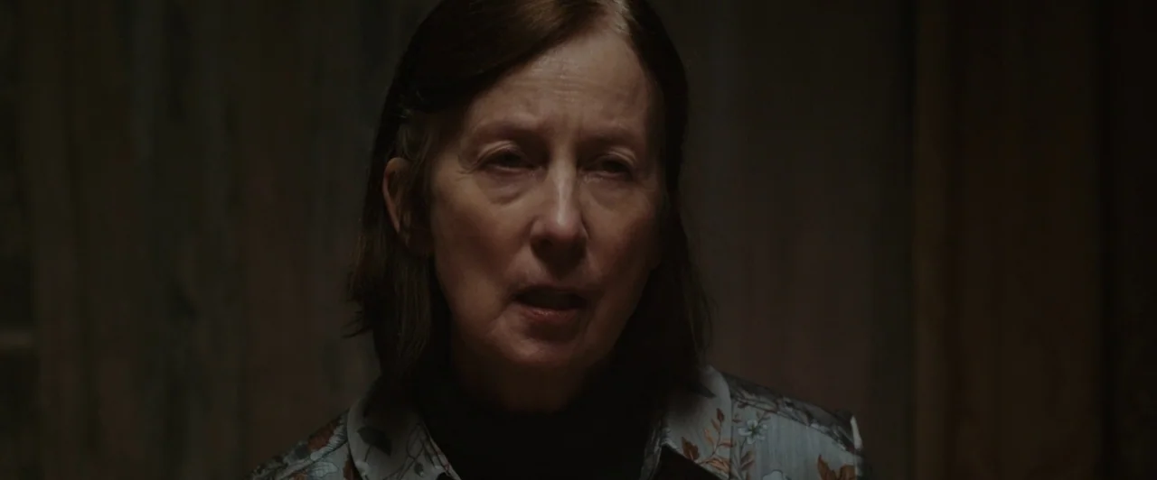 You Are Not My Mother (2021) movie still: closeup — A dimly lit indoor space with indistinct dark walls in the background. A woman in a floral shirt…