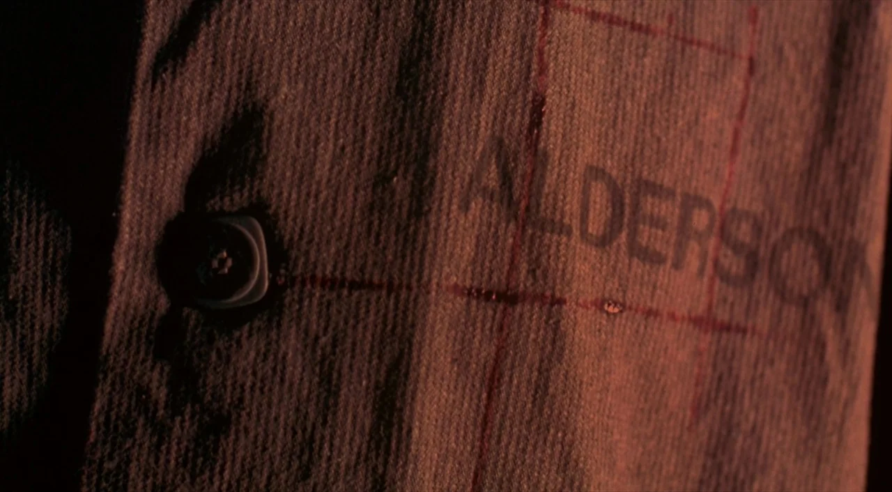 Cube (1997) movie still: closeup — A close-up of a coarse, red textile with a button and a stamped name. The name ALDERSON is clearly printed in…
