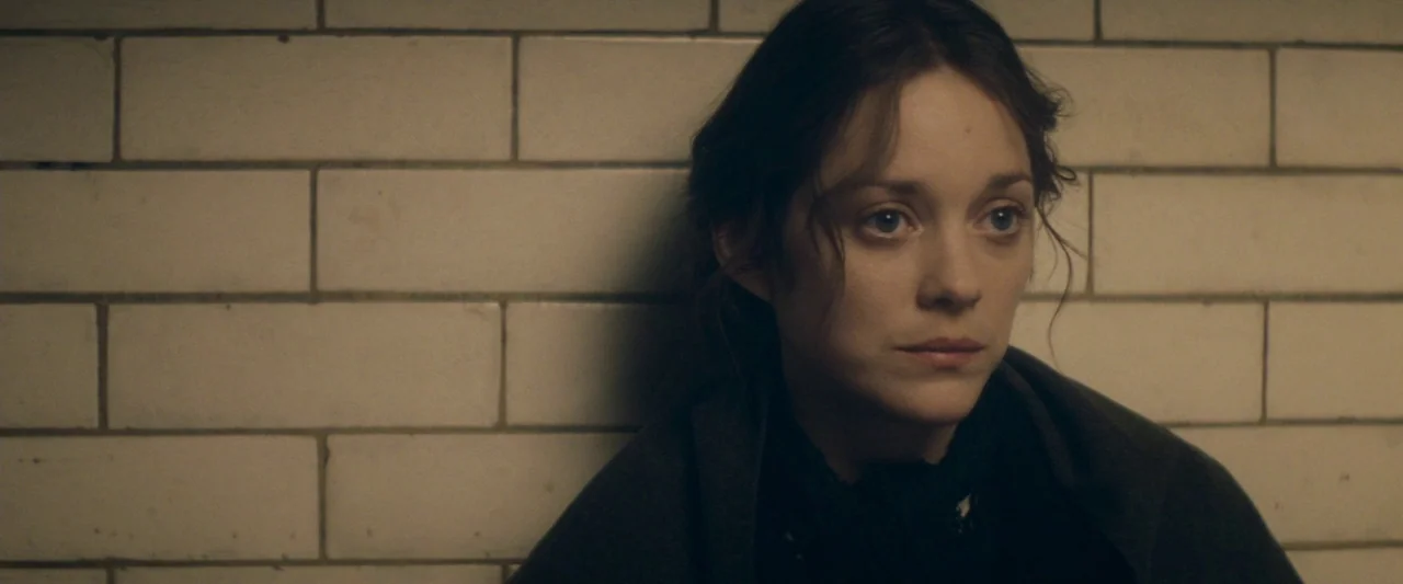 The Immigrant (2013) movie still: closeup — A woman leans against a white tiled wall in a dimly lit, austere space. She is positioned slightly to…