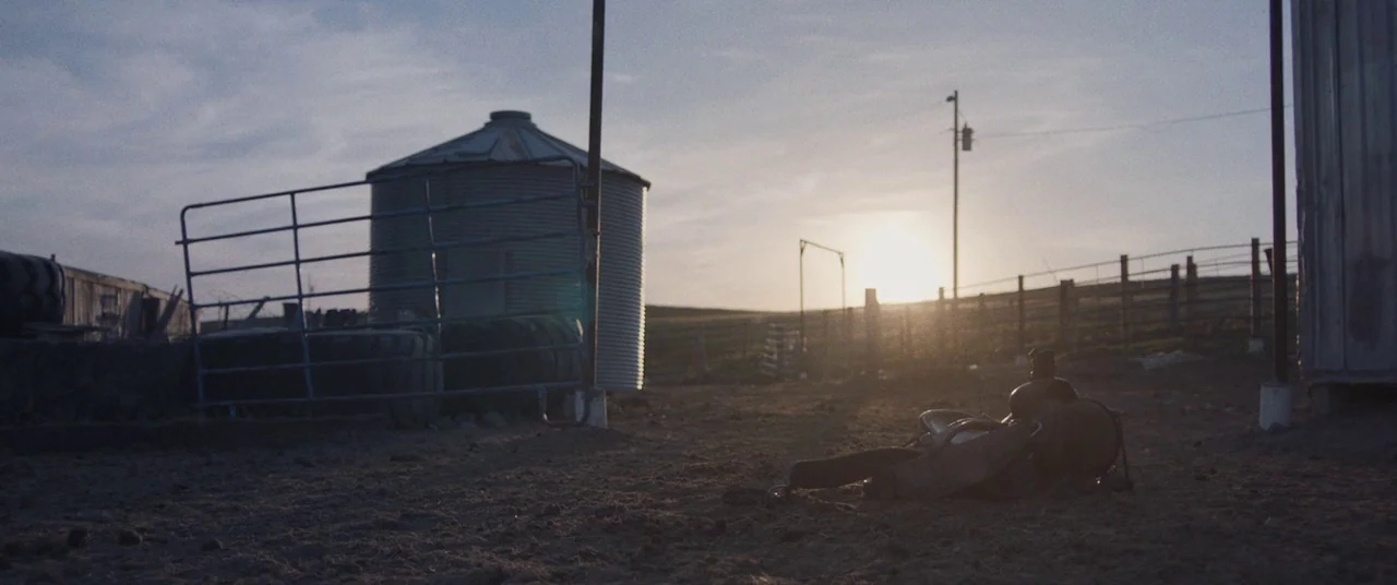 The Rider (2017) movie still: wide — A rural farmyard at sunset features a metal silo and a blue metal gate. A single western saddle rests on the…