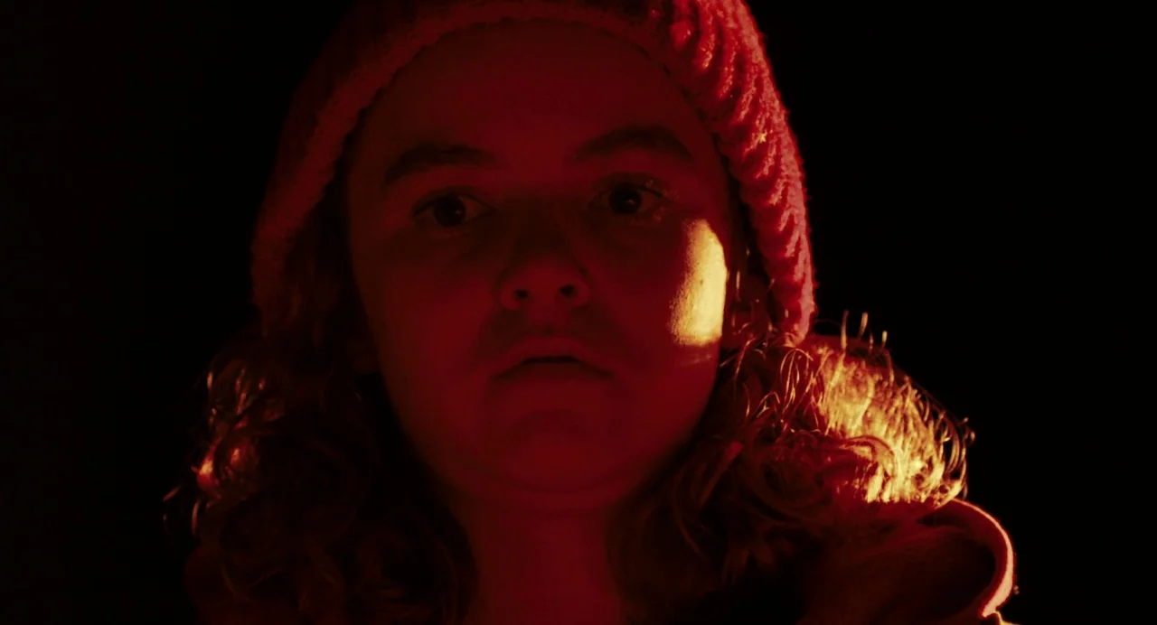 Blow The Man Down (2019) movie still: closeup — A dark space surrounds a person whose face is illuminated by bright red light. The person wearing a…