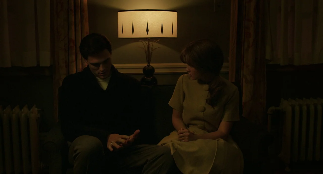 Priscilla (2023) movie still: medium — A dimly lit room with muted walls and a central table lamp on a sofa. A man and a woman sit together in the…