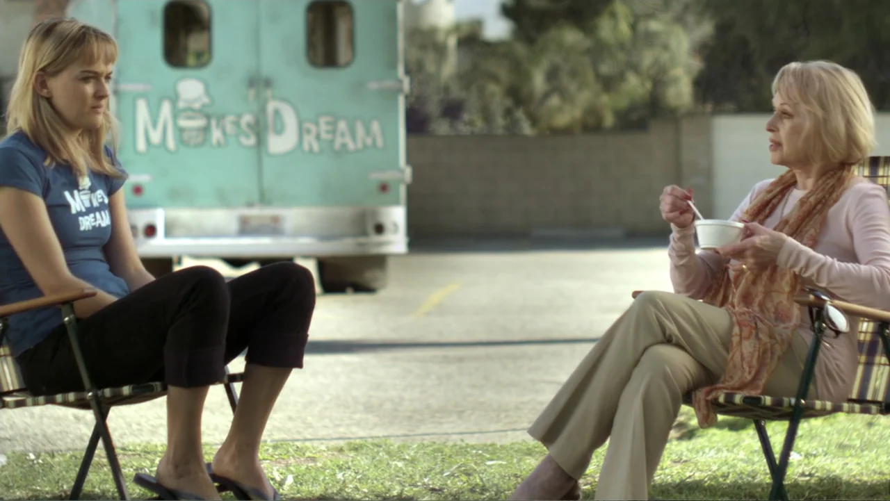 Free Samples (2013) movie still: medium — A sunny parking lot contains a parked light blue vintage ice cream truck. Two women sit in folding chairs,…