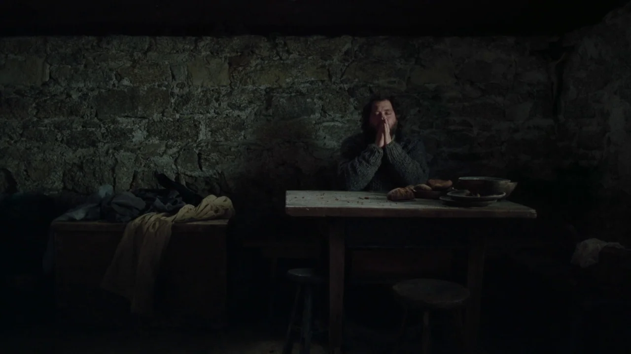 The Devil’s Bath (2024) movie still: wide — A dimly lit room features rough stone walls and basic wooden furniture. A man in a thick sweater sits at…