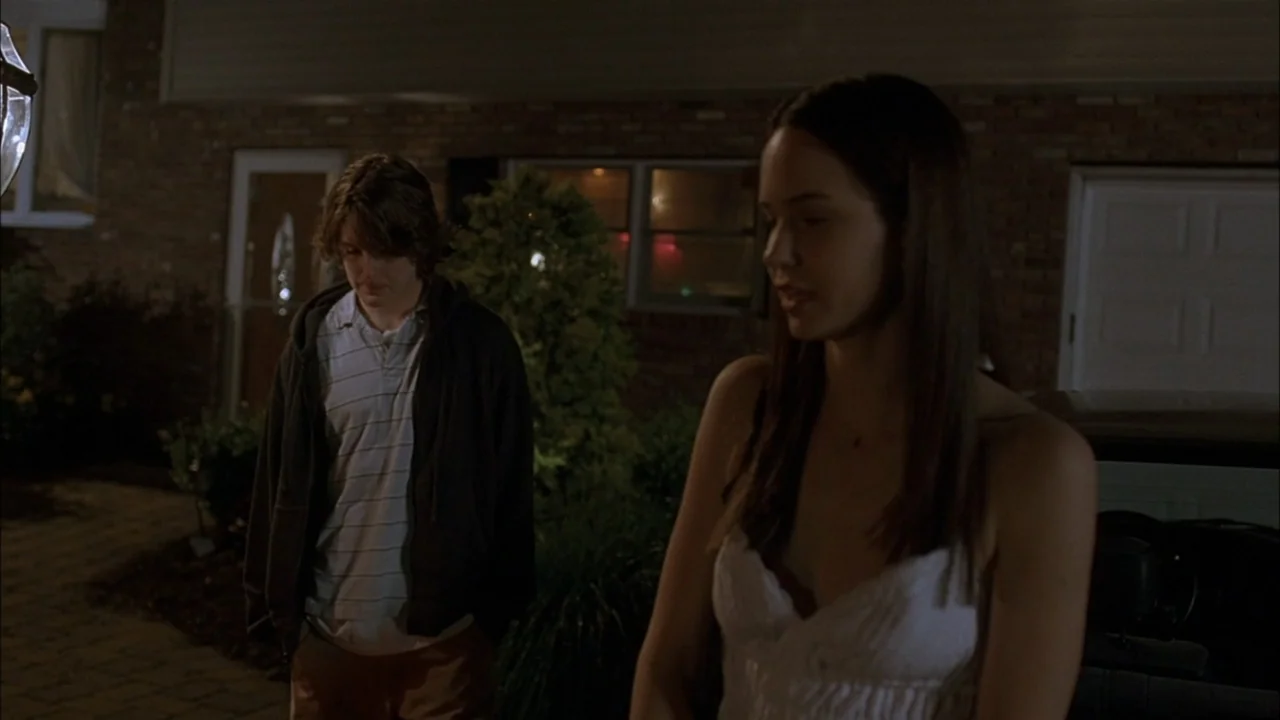 The Babysitters (2008) movie still: medium — A paved residential driveway at night with a brick house facade visible in the background. A young…