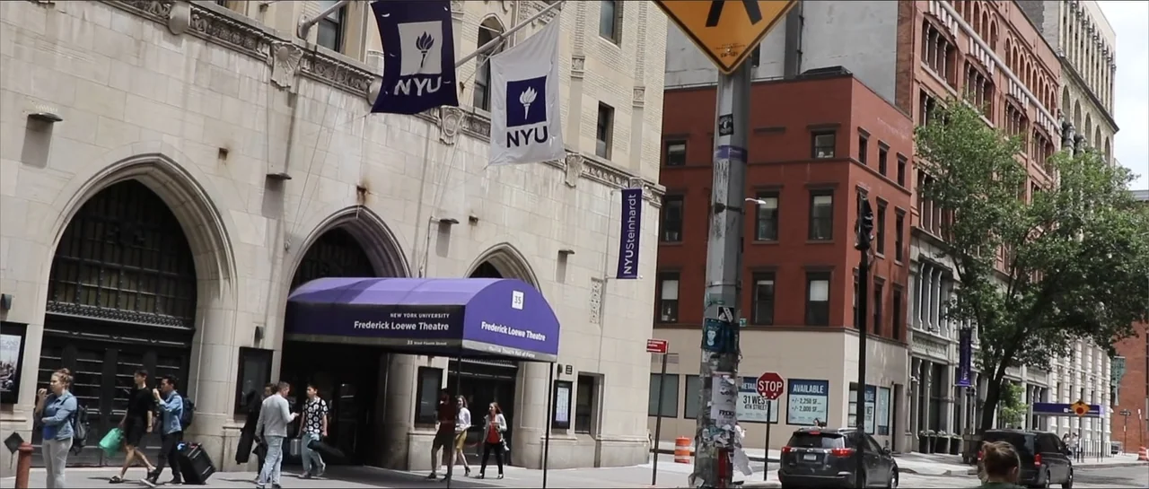 Square One (2019) movie still: wide — A stone theater building with purple awnings and NYU flags sits on a city street. Several people walk on the…