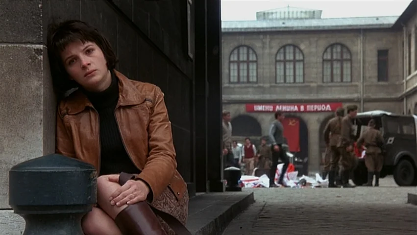 The Unbearable Lightness of Being (1988) movie still: medium — A paved town square with stone buildings and a banner hanging over an archway. A…