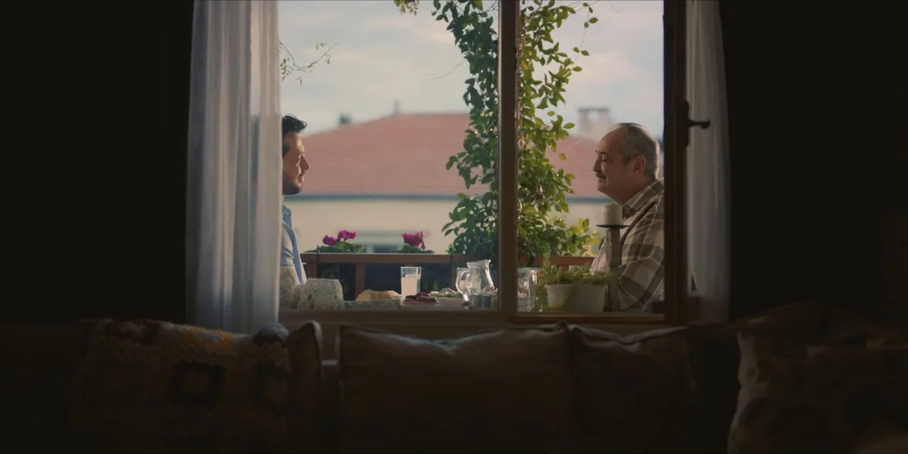 Kill Me If You Dare (2019) movie still: medium — Two men sit at a small table on a sunlit balcony with flowering plants. The men engage in a…