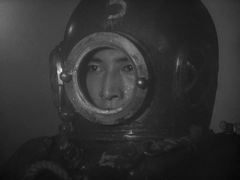 Godzilla (1954) (1954) movie still: closeup — A close-up of a person wearing a heavy, vintage-style brass or steel diving helmet. The subject stares…