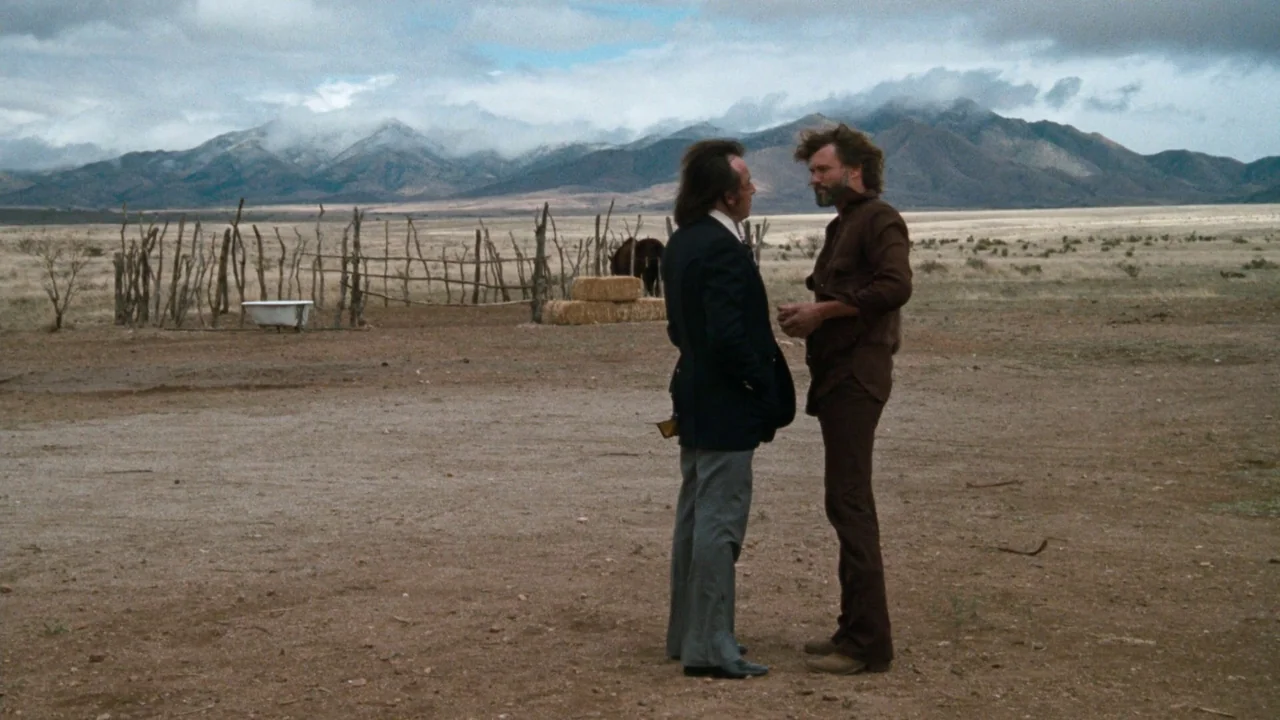 A Star Is Born (1976) movie still: fullbody — A vast, dry desert landscape features a wooden fence and distant snow-capped mountains. Two men stand…