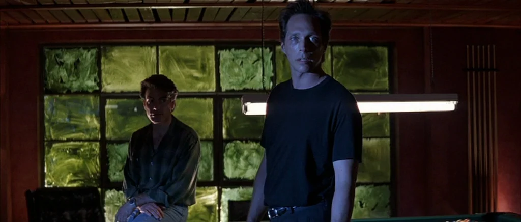 The Underneath (1995) movie still: medium — A dimly lit room features a large window with paint-smeared green glass panels. Two men stand near a…