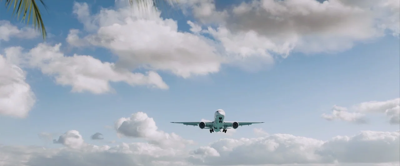 Beautiful Wedding (2024) movie still: wide — A commercial jet airliner approaches in the center of a wide, cloud-filled blue sky. Green palm tree…