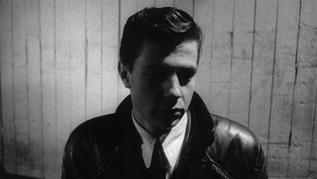 Who’s That Knocking At My Door (1967) movie still: closeup — A plain wall with vertical wooden paneling serves as the backdrop. A young man stands…