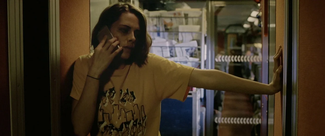 Personal Shopper (2016) movie still: medium — A narrow corridor inside a modern train with wood-paneled walls and metal doorways. A woman stands in…