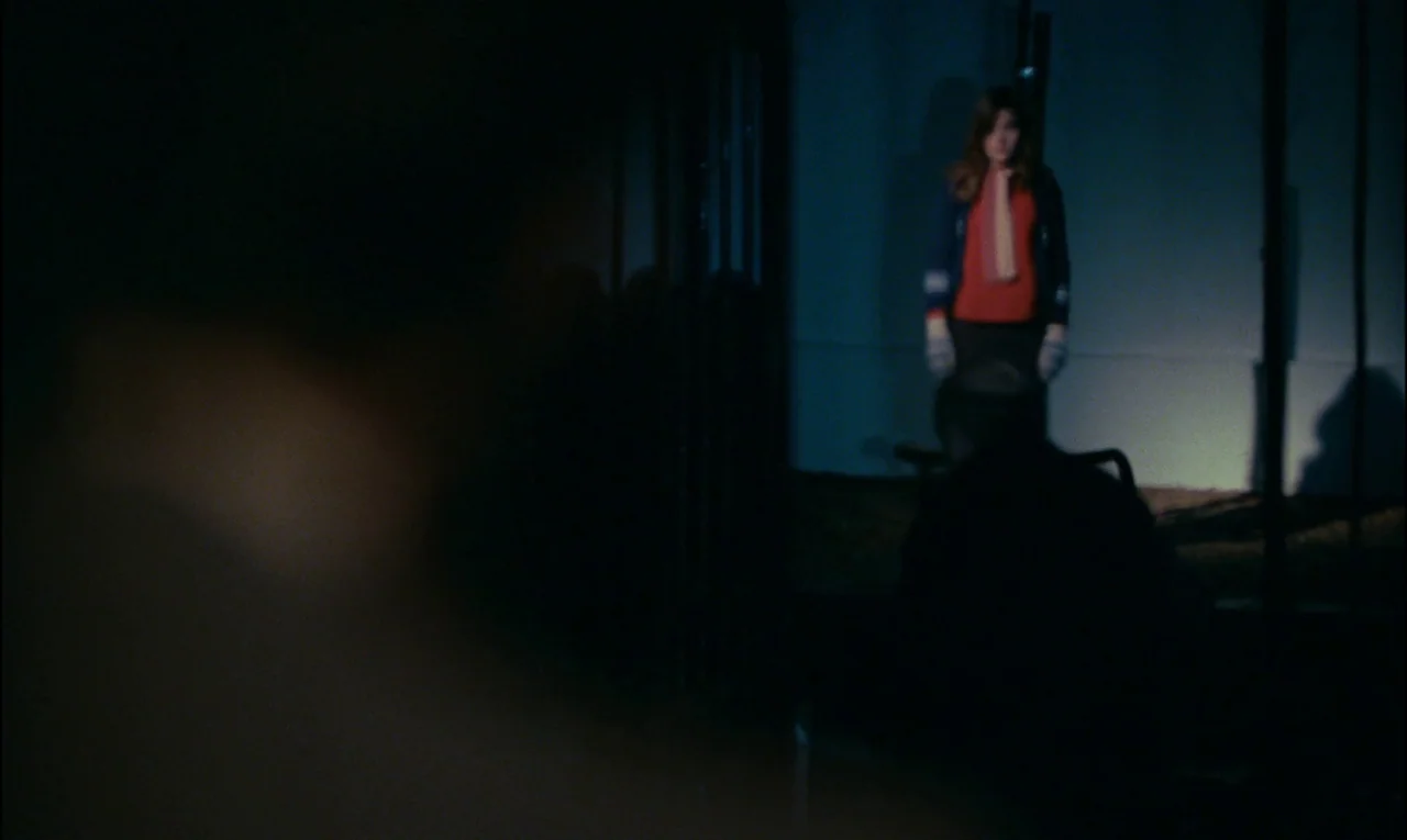 Serie Noire (1979) movie still: wide — A dimly lit exterior nighttime environment features a stark white wall behind a woman. The woman stands alone…