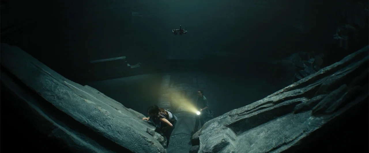 Black Adam (2022) movie still: wide — A vast underground stone chamber features jagged rock walls and ancient architectural geometric carvings. Two…
