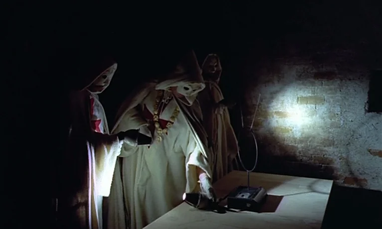 Nuits Rouges (1974) movie still: medium — A dark, unfinished basement with rough brick walls and a flat wooden table. Three figures in cream-colored…