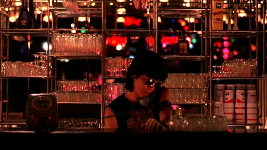 The Terrorizers (1986) movie still: medium — A dimly lit bar counter features metal shelving filled with numerous glass tumblers. A person wearing…
