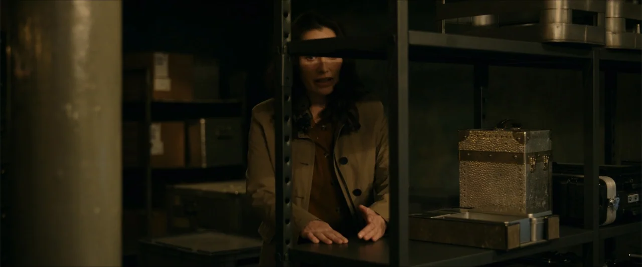 Gunpowder Milkshake (2021) movie still: medium — A dark, cluttered storage room contains metal shelving units and various opaque crates. A woman in…