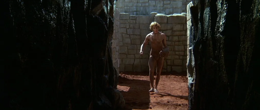 Fellini – Satyricon (1969) movie still: fullbody — A stone-walled labyrinth features narrow passages with red-brown dirt floors. A muscular man…