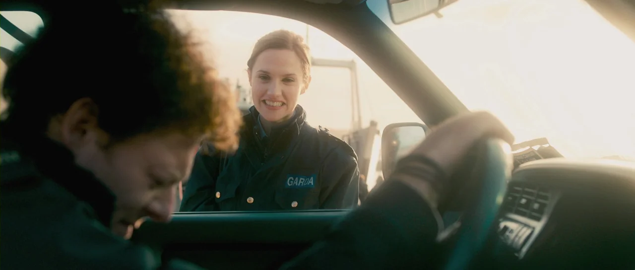 Grabbers (2012) movie still: medium — A female Garda officer stands outside a car window on a bright, hazy day. She smiles down at the driver whose…