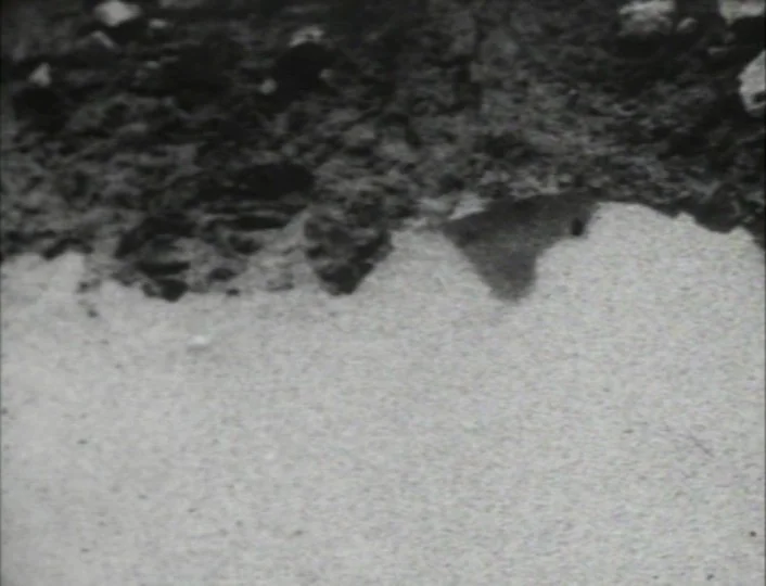 Begotten (1989) movie still: establishing — A grainy black and white view of the uneven, dusty lunar surface. The terrain consists of dark volcanic…