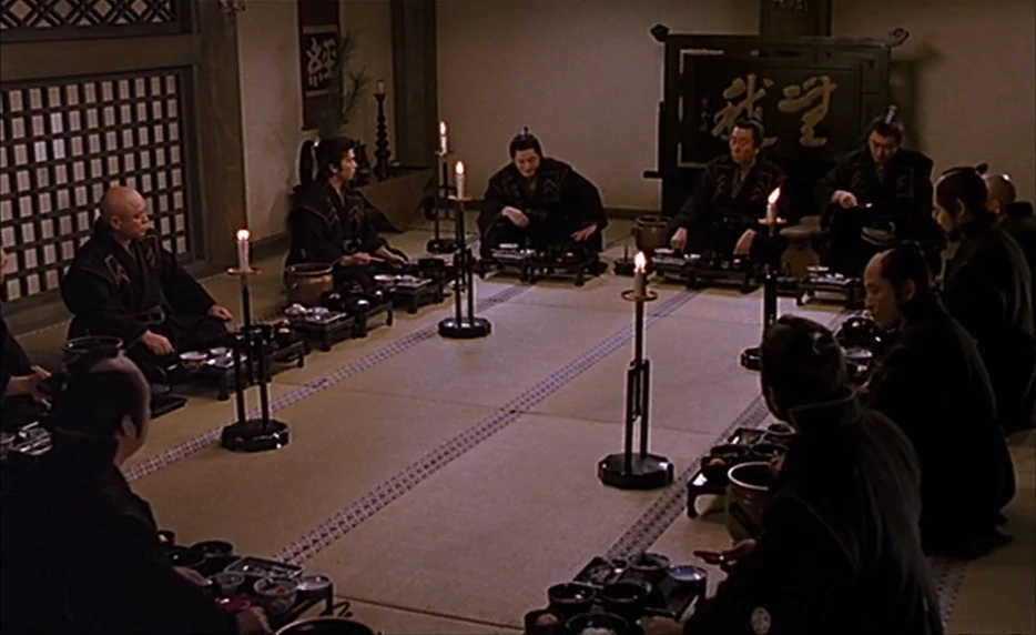 Gohatto (1999) movie still: wide — A square room with tatami mats holds a group of men in traditional Japanese robes. They sit at low tables…