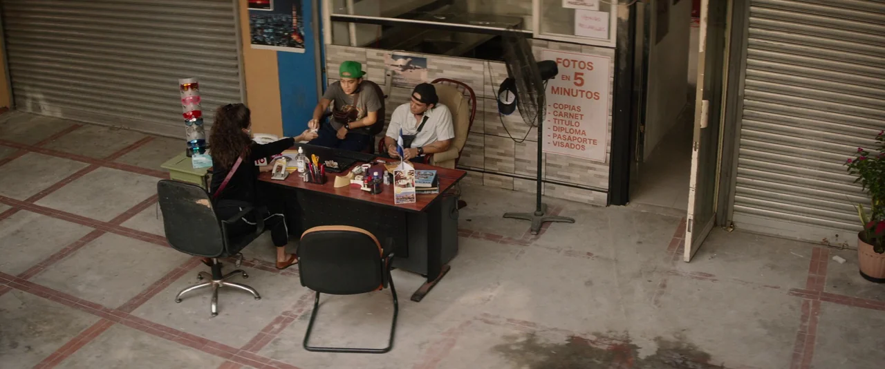 Stars At Noon (2022) movie still: wide — A small business office features tiled walls and a sign listing photo services. Three people sit at a…