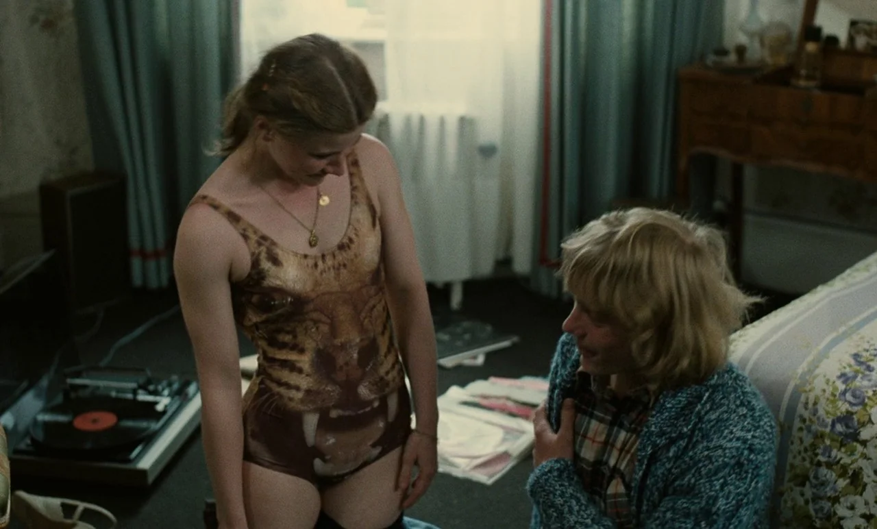 Graduate First (1978) movie still: medium — A cluttered 1970s room with stacks of vinyl records on the carpeted floor. A woman wearing a tiger…