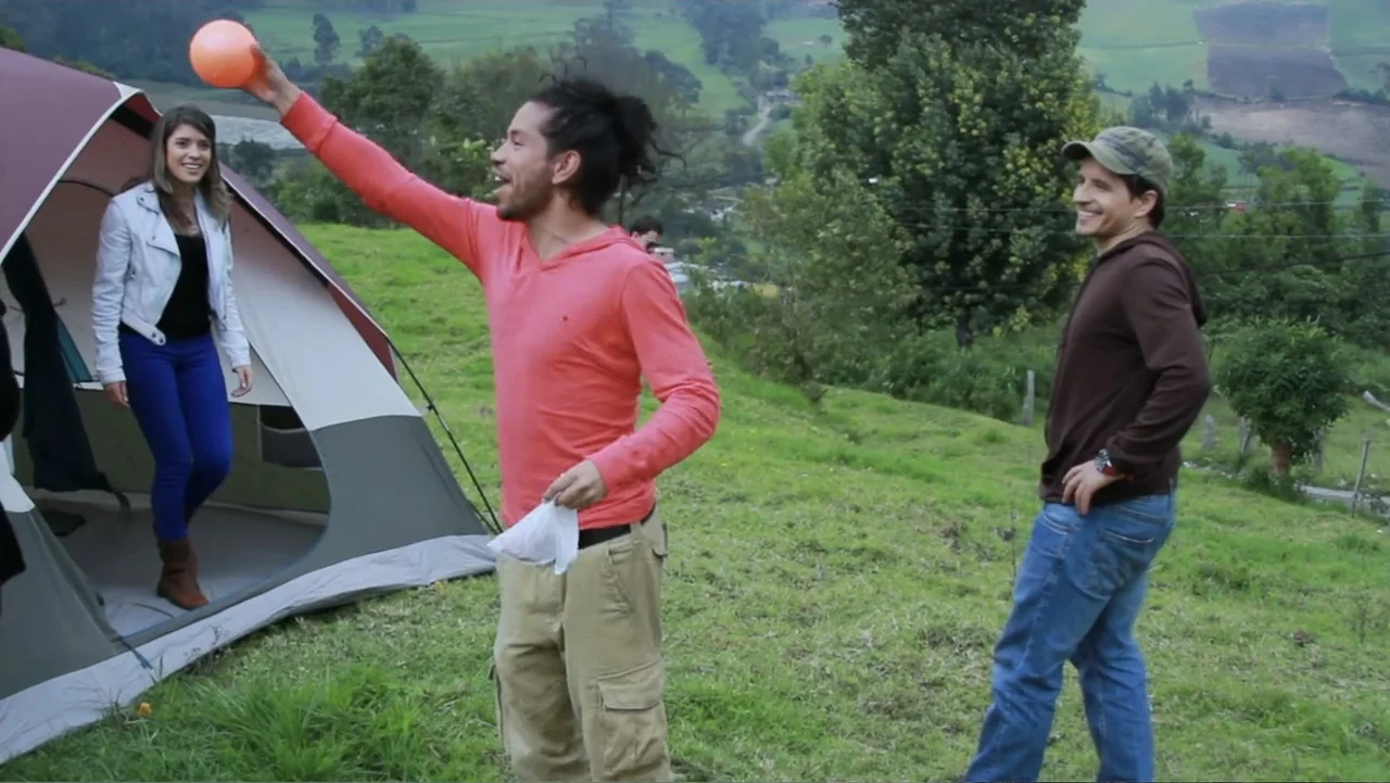Aquel Septiembre (2018) movie still: fullbody — A green grassy hillside features a camping tent pitched near three young adults. The man in the…